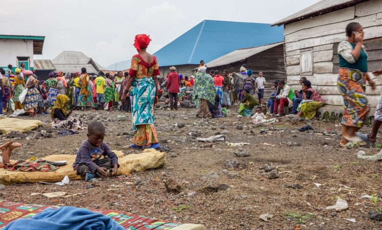 People gather in a village with colorful clothing. Children play on the ground, while others converse near rustic houses.