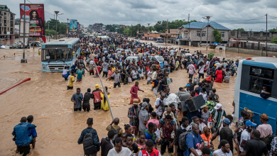 Crowd wades through floodwaters on a congested street, with buses and vehicles partially submerged under a cloudy sky.