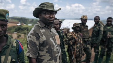 A group of soldiers in camouflage uniforms stand together outdoors, with one in a hat looking towards the camera.