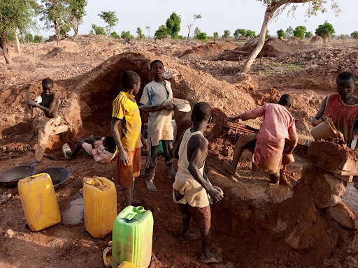 Children working in a rural area with tools and water containers, surrounded by dirt and trees.
