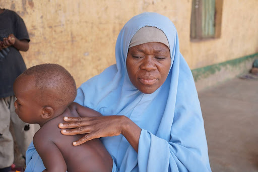 A woman in a blue hijab holds a young child, pressing on the child's back, outside a rustic building.